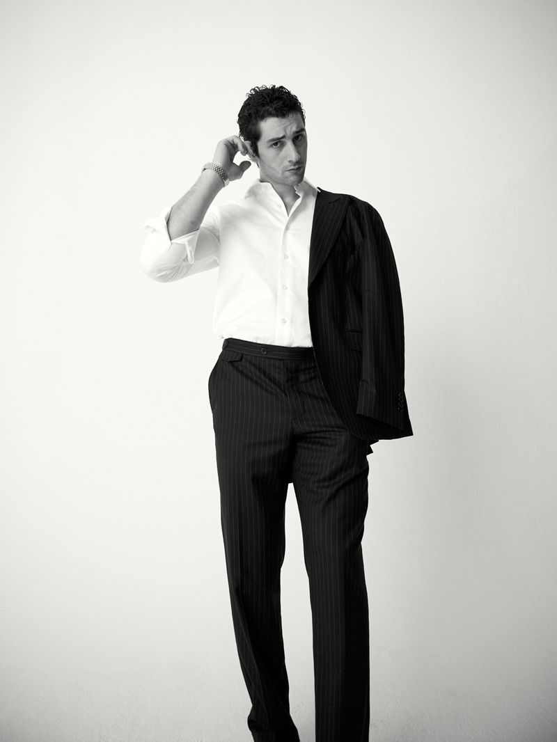 Monochrome portrait of a man in a partially buttoned white shirt and suit trousers with a jacket draped over his shoulder.