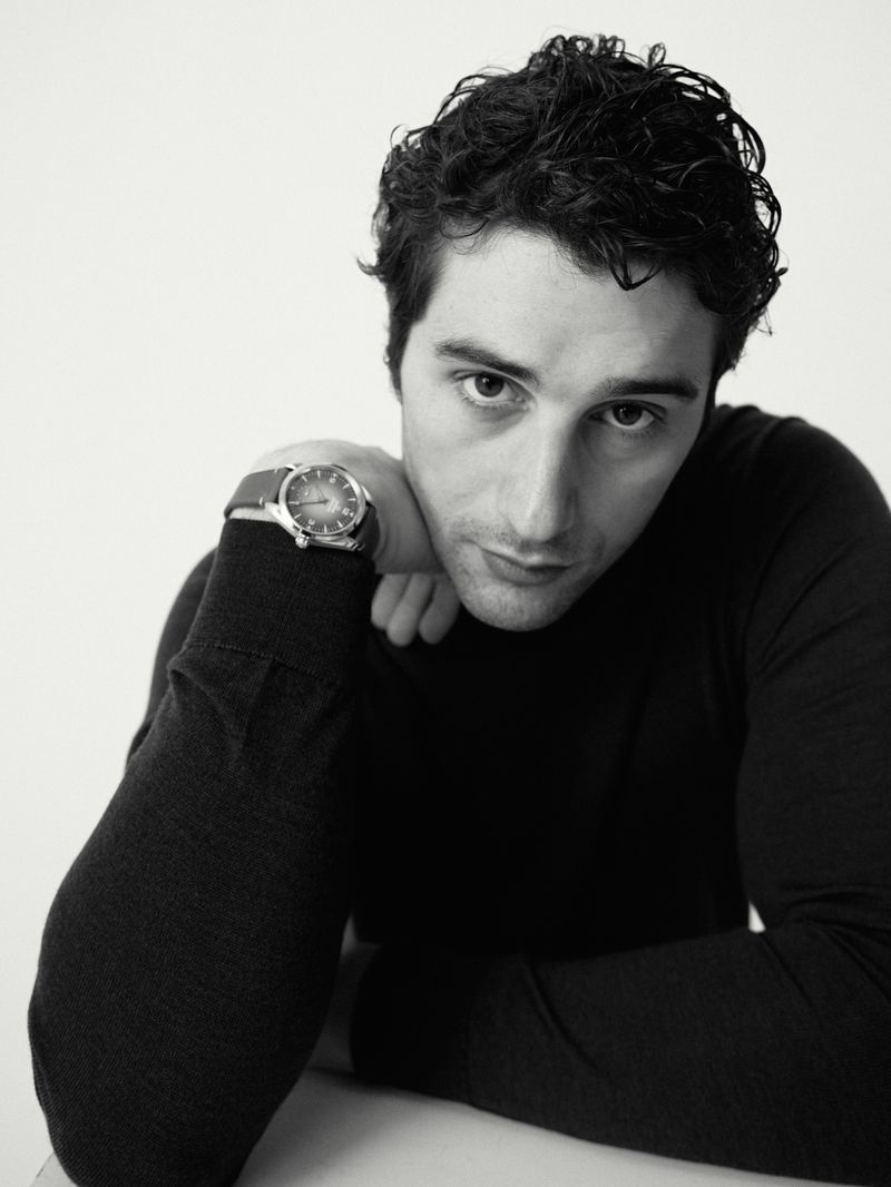 Man with dark wavy hair resting his chin on his arm, wearing a black knit sweater and wristwatch against a plain white studio backdrop.