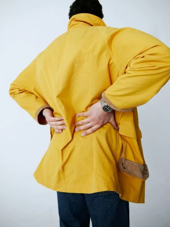 Adult male wearing a yellow jacket, seen from the back. Jacket has brown accents and large pockets. Man also wears blue jeans.