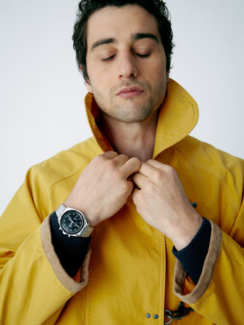 Adult male in a yellow coat, eyes closed, adjusting his collar, wearing a stainless-steel wristwatch. Indoor studio shot with soft lighting.