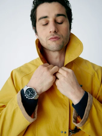 Adult male in a yellow coat, eyes closed, adjusting his collar, wearing a stainless-steel wristwatch. Indoor studio shot with soft lighting.