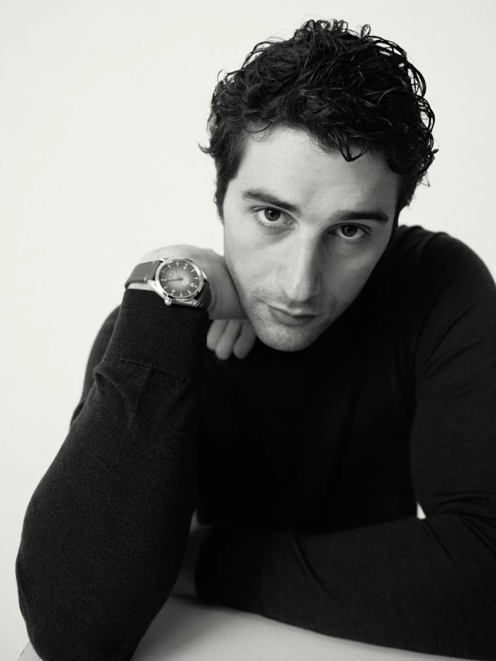 Man with dark wavy hair resting his chin on his arm, wearing a black knit sweater and wristwatch against a plain white studio backdrop.