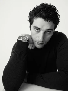 Man with dark wavy hair resting his chin on his arm, wearing a black knit sweater and wristwatch against a plain white studio backdrop.