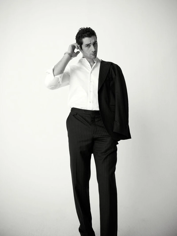 Man in white shirt and pinstripe suit trousers touching his hair against a plain white background for Risbel Magazine editorial