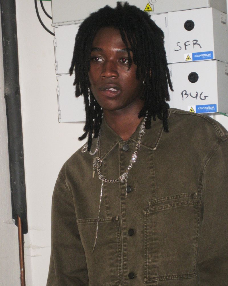 Portrait of a young adult with dreadlocks wearing a green jacket and silver necklace, standing indoors in front of white boxes.