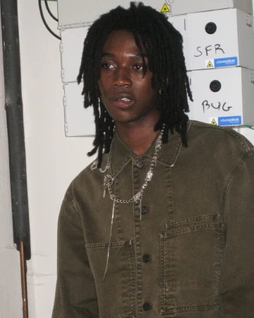 Portrait of a young adult with dreadlocks wearing a green jacket and silver necklace, standing indoors in front of white boxes.