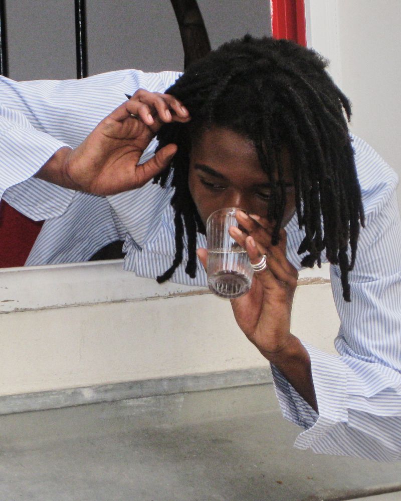 Adult drinks from glass, wearing a striped shirt; black dreadlocks. The backdrop shows a combination of white, grey, and red colours.
