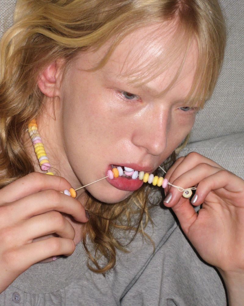 A fair-skinned young adult woman with blonde hair poses with candy necklace in her mouth and hands indoors, showcasing a playful fashion statement.