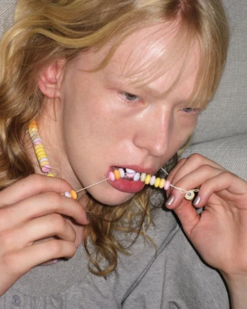 A fair-skinned young adult woman with blonde hair poses with candy necklace in her mouth and hands indoors, showcasing a playful fashion statement.
