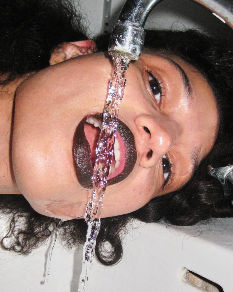 Upside-down portrait of a woman with dark lipstick drinking water from a chrome faucet in a white sink, eye-level shot.