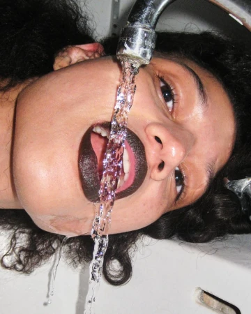 Upside-down portrait of a woman with dark lipstick drinking water from a chrome faucet in a white sink, eye-level shot.