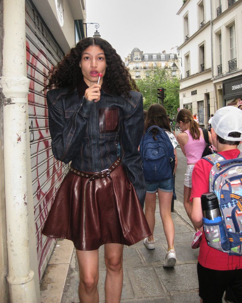 Fashionable woman in denim jacket and brown leather skirt with a lollipop standing on a city street with people and buildings.