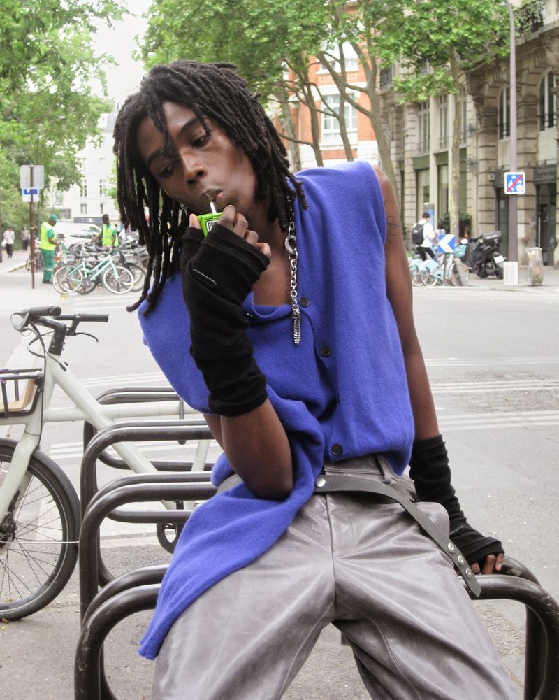 Young adult stands on a city street, drinking from a juice box. Wears a blue vest, gray pants, and fingerless gloves, accessorized with chains.