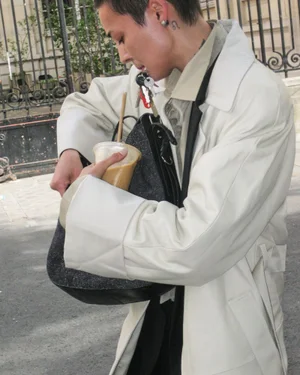Model in an oversized off-white coat holding an iced coffee and searching through a black shoulder bag outdoors.