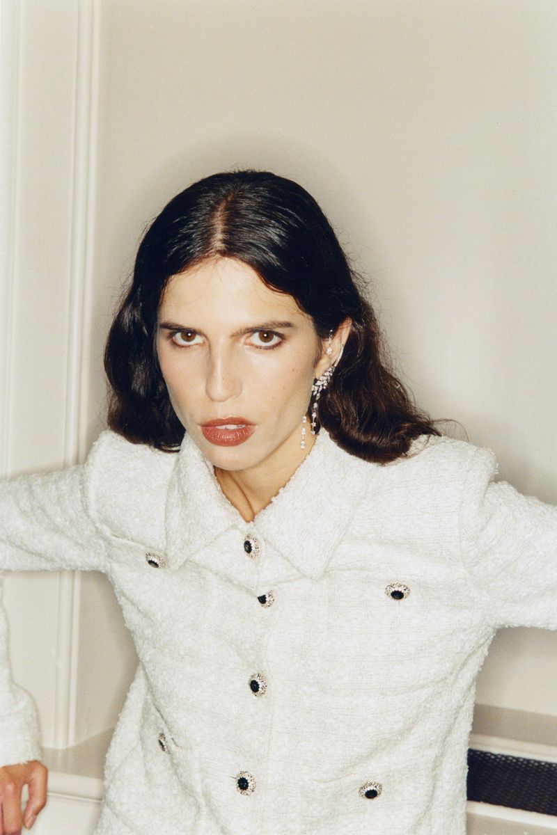 Woman in white textured jacket with dark hair and statement earrings posing indoors against a neutral wall with architectural details.