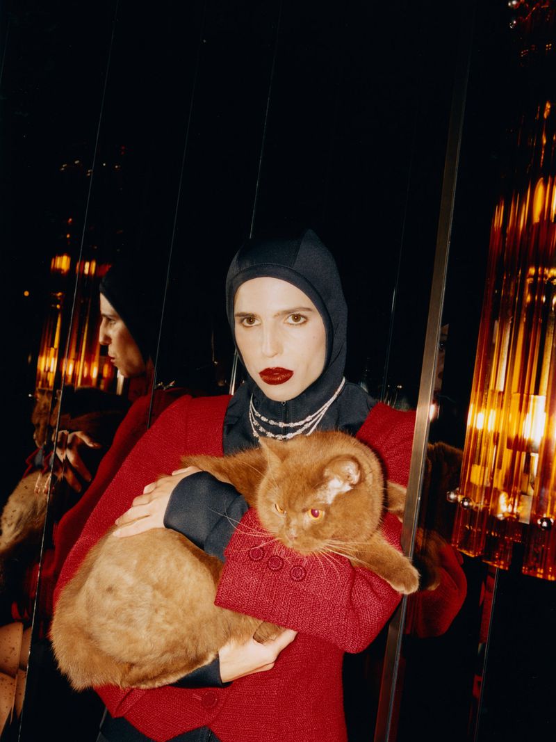 A woman with fair skin, in a dark hooded top, red jacket, and pearl necklace, holds a ginger cat in an indoor, dark environment.