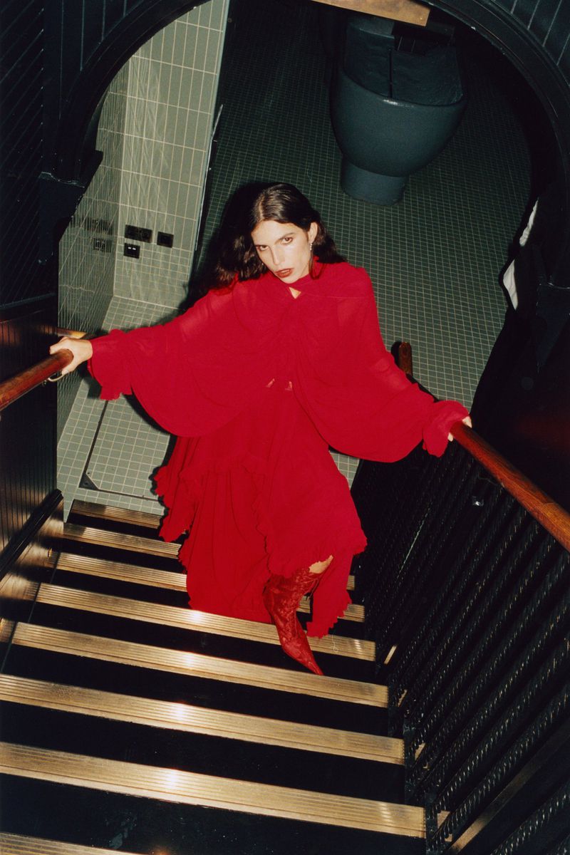 Fashionable woman in a bright red dress and matching boots poses on a staircase inside a building with a tiled wall and restroom.