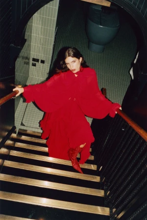 Fashionable woman in a bright red dress and matching boots poses on a staircase inside a building with a tiled wall and restroom.