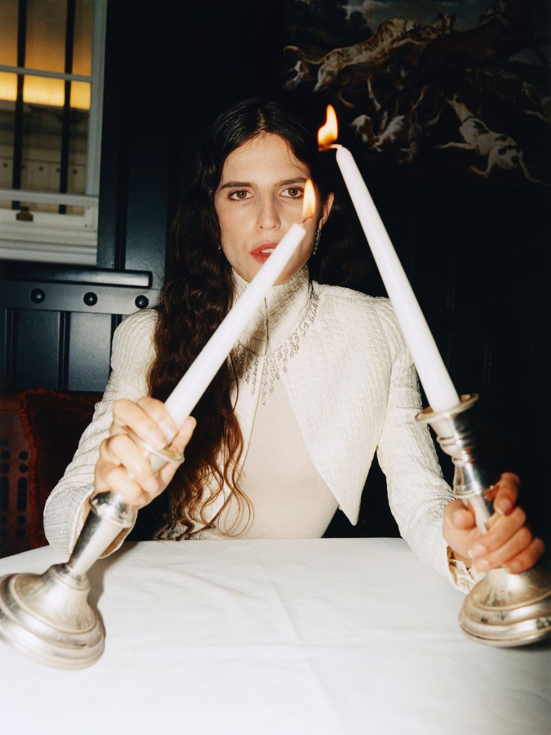 Brunette woman in a cream-colored jacket holds lit candles at a table. Moody indoor setting with a dark background and formal attire.