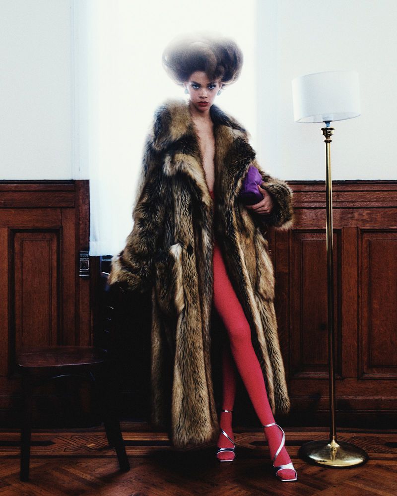 Elegant adult female fashion model in a long fur coat, pink stockings, silver shoes, and chair and lamp in an indoor setting.