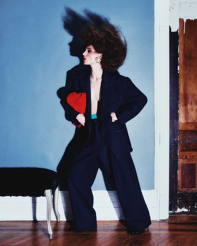 Stylish woman in dark suit with red clutch poses in front of a blue wall, casting a shadow of her voluminous hair in a fashion shot.