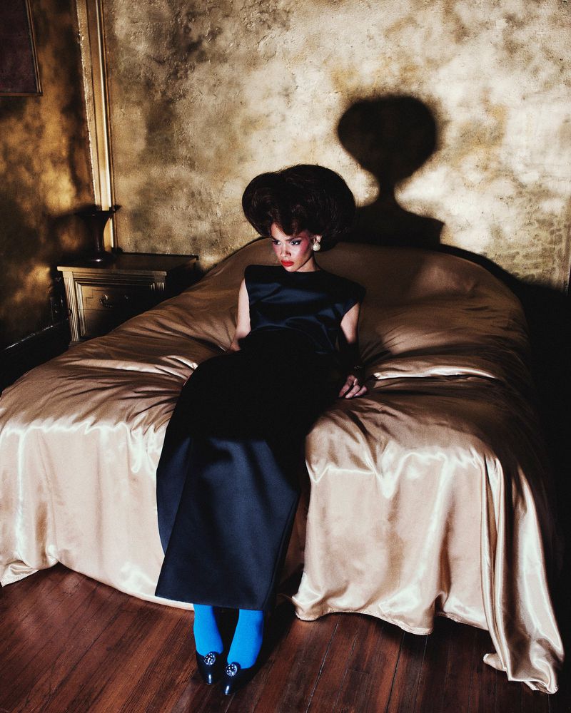 A woman with an afro is sitting on a bed in a dramatic pose. She wears a black dress, bright blue tights, and high-heeled shoes.