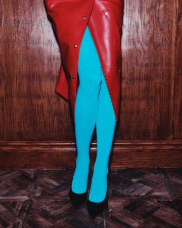 Model wearing a red leather wrap skirt, bright turquoise tights, and black pointed-toe high heels against a dark wood-paneled wall.