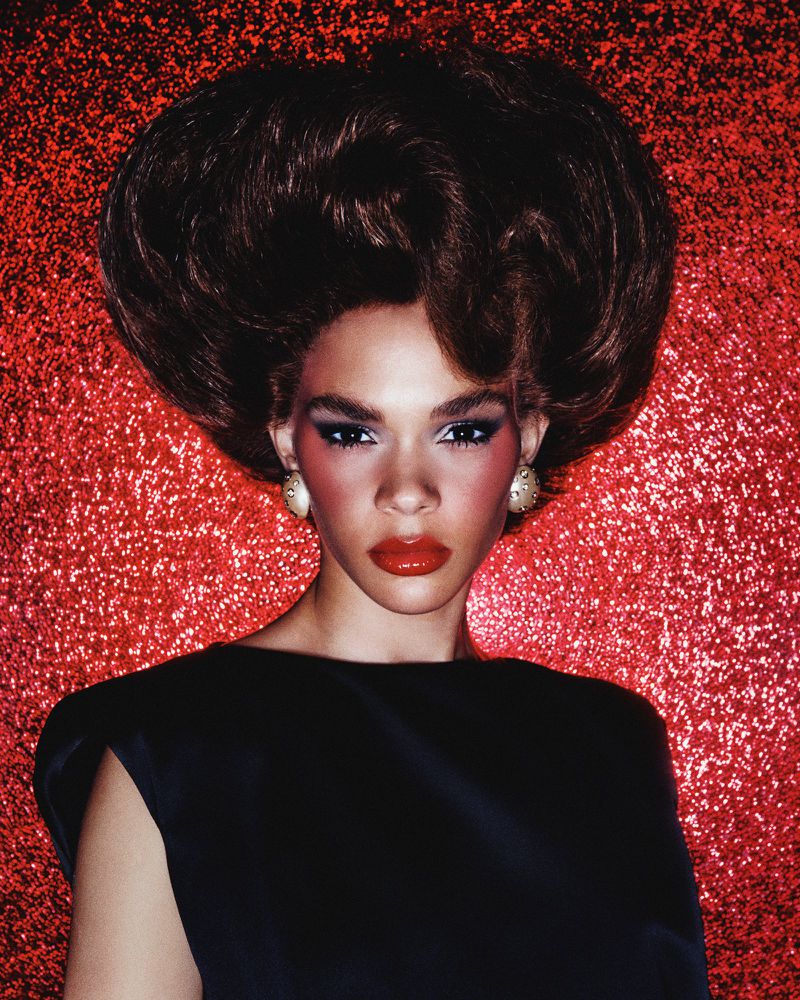 Portrait of a woman with dark hair in a beehive style, wearing a black top against a textured, glittery red backdrop in a studio.