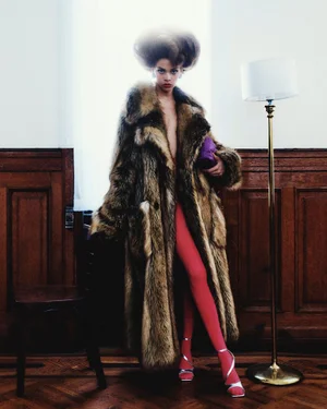 Model in voluminous fur coat and pink tights standing in an interior with dark wood paneling and a floor lamp.