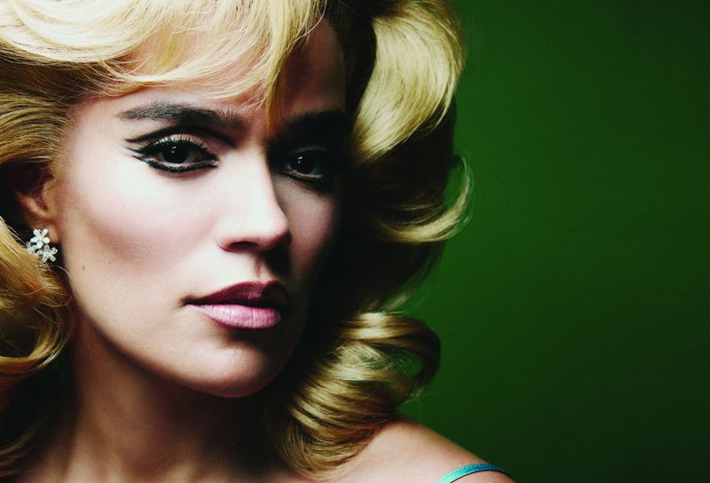 Close-up portrait of Karol G with vintage blonde voluminous hair and heavy winged eyeliner against a dark green backdrop