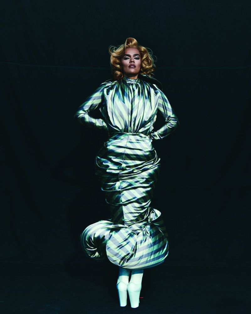 Blonde woman posing in a silver metallic striped high fashion gown with dramatic draping and white pointe shoes on a dark background.
