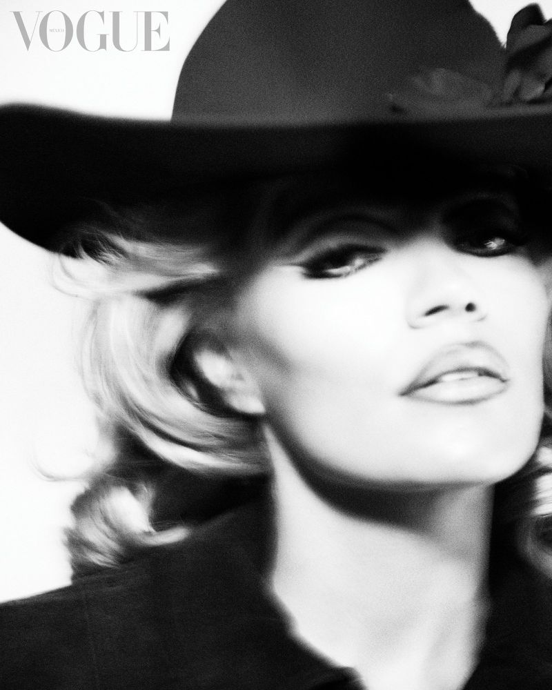 Black and white close-up portrait of Karol G wearing a dramatic wide-brimmed black hat and dark coat.
