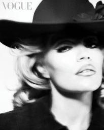 Black and white close-up portrait of Karol G wearing a dramatic wide-brimmed black hat and dark coat.