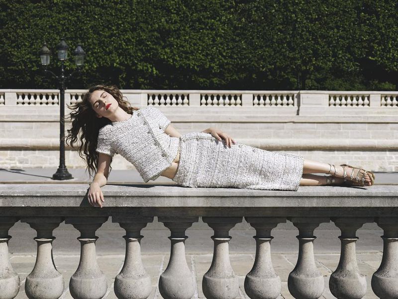 A woman in a tweed suit lies on a stone wall with a green hedge and lamppost in the background, in a fashion advertisement-style outdoor shot.