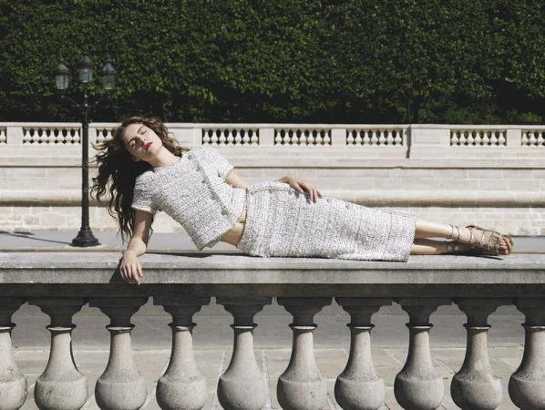 A woman in a tweed suit lies on a stone wall with a green hedge and lamppost in the background, in a fashion advertisement-style outdoor shot.