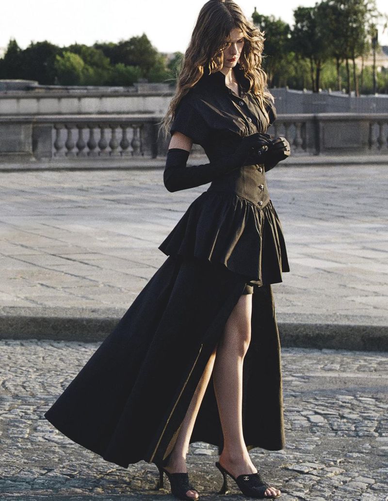 A woman in a black dress with gloves and heels poses on cobblestones in a high-fashion outdoor shot with a city background.