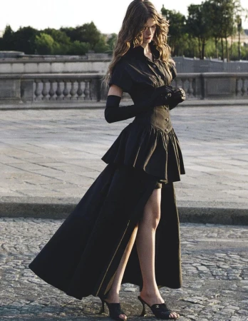 A woman in a black dress with gloves and heels poses on cobblestones in a high-fashion outdoor shot with a city background.