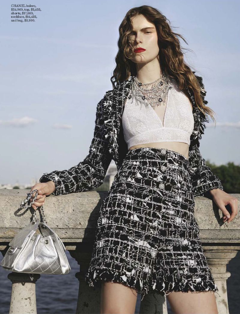 Woman in tweed jacket and skirt, silver bag, and necklace poses outside on a stone bridge with water and sky in the background.