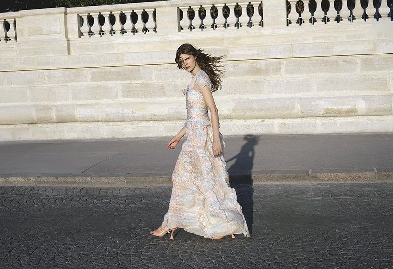 Woman in a flowing floral dress walking on a cobblestone street next to a stone wall under natural lighting, fashion photography.