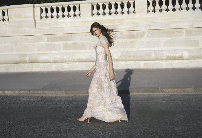 Woman in a flowing floral dress walking on a cobblestone street next to a stone wall under natural lighting, fashion photography.