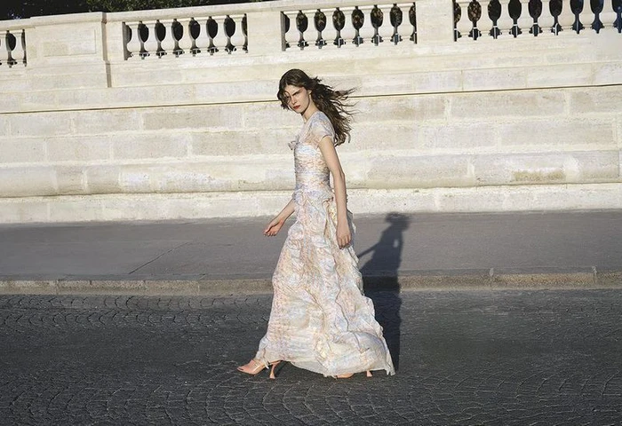 Model Anna Robinson walking along a street wearing a pastel tiered gown in front of a stone balustrade.