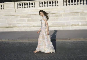 Model Anna Robinson walking along a street wearing a pastel tiered gown in front of a stone balustrade.