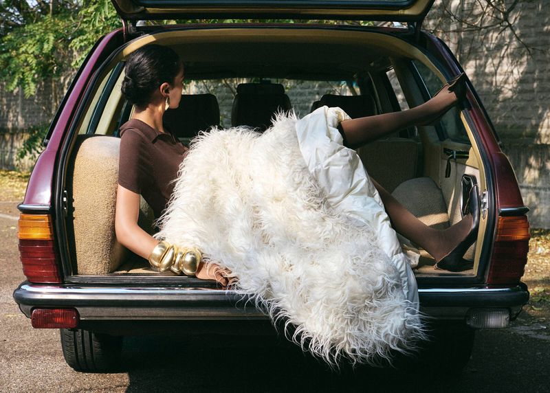 Woman poses in vintage car trunk with fur blanket, wearing a brown shirt and gold jewelry, and black high heels, outside near a brick wall.