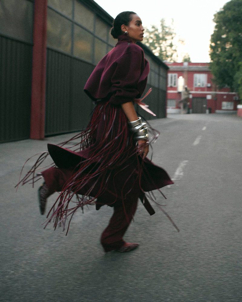 Fashionable woman in burgundy outfit with fringe running on paved road alongside industrial building with closed metal doors and red trim.