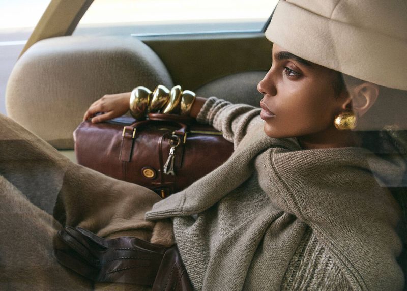 Adult female wearing a hat and gold bracelets, posing inside of a vehicle with a leather travel bag, wearing a sweater and looking away.
