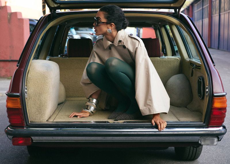 Stylish woman poses in open trunk of burgundy station wagon wearing trench coat, sunglasses, green tights and silver jewelry.