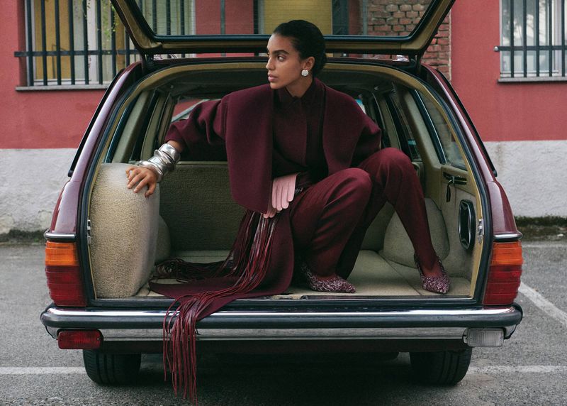 Woman in burgundy outfit poses inside car trunk in urban setting, accessorized with silver cuff, against brick building background.