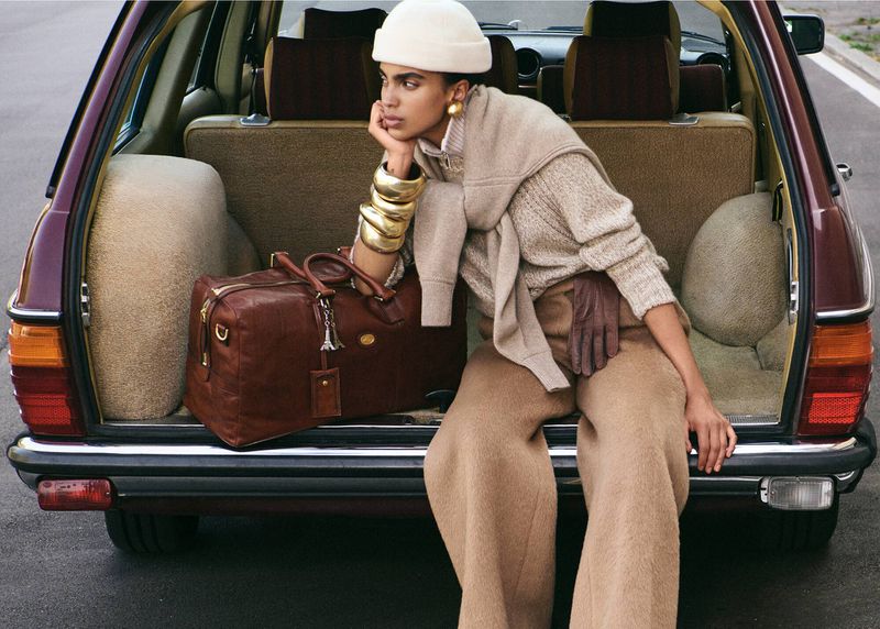 Stylish woman in beige outfit poses in classic car trunk with leather duffel bag and gold bracelets, waiting for travel.