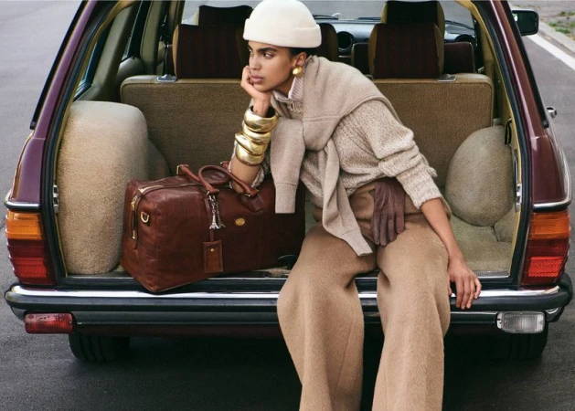 Stylish woman in beige outfit poses in classic car trunk with leather duffel bag and gold bracelets, waiting for travel.