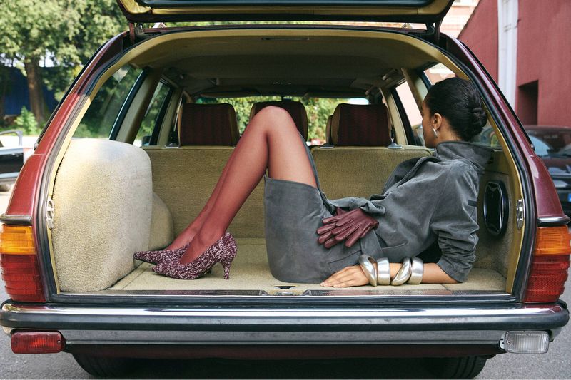Adult woman lies in the trunk of an older model car, wearing a gray skirt, gray jacket, sheer red stockings, and glittery high heel shoes.